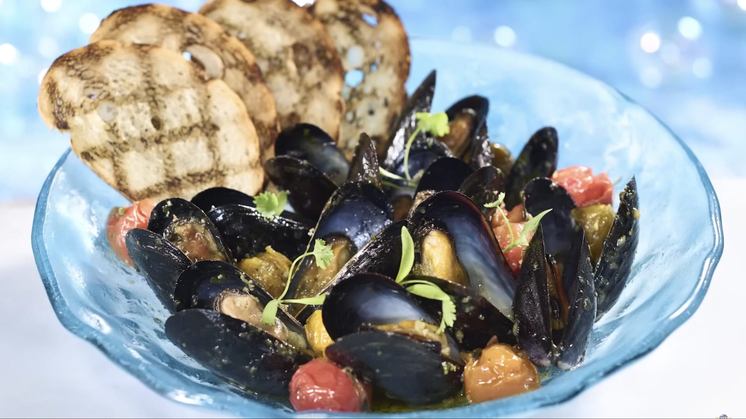 A bowl of mussels with cherry tomatoes and herbs, served with two slices of toasted bread, showcases the intricate details of exquisite food and beverage perfection.